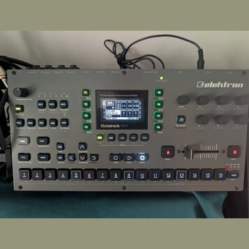 Stříbrný sampler s nápisem Elektron a Octatrack MKII. Zařízení obsahuje displej, několik ovládacích knoflíků, tlačítka s různými funkcemi a v dolní č…