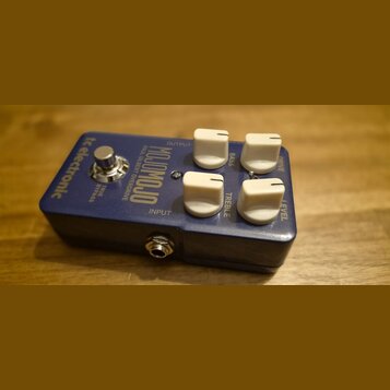 Modrý kytarový efekt s bílými ovládacími knoflíky. Na šasi jsou nápisy tc electronic, MOJOMOJO a PAUL GILBERT OVERDRIVE. Viditelné popisky u knoflíků…