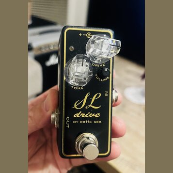 Černý kytarový efekt s zlatým vnitřním rámem a nápisem SL drive BY xotic usa. Na horní straně jsou tři ovládací prvky: otočný knoflík TONE, otočný kn…