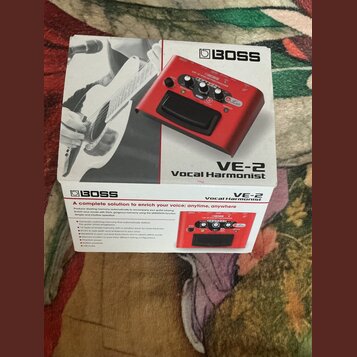 Krabice s červeným vokálovým procesorem značky BOSS, model VE-2 Vocal Harmonist. Na krabici je produktová fotografie zařízení a černobílá fotografie…