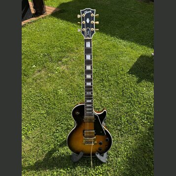Elektrická kytara s tónem těla v barvě sunburst a černým pickguardem. Na hlavě je viditelné logo Gibson a nápis Custom. Hриф má vložky z perlemi a na…