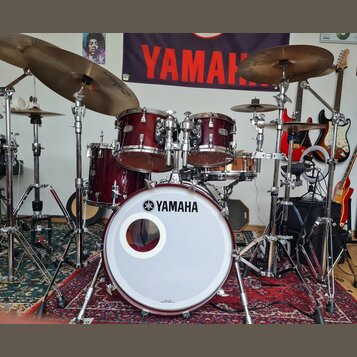Bicí souprava v tmavě červené barvě s kovovým hardwarem. Na bílém předním z koženě basového bubnu je čitelné logo a nápis YAMAHA. V pozadí visí černý…