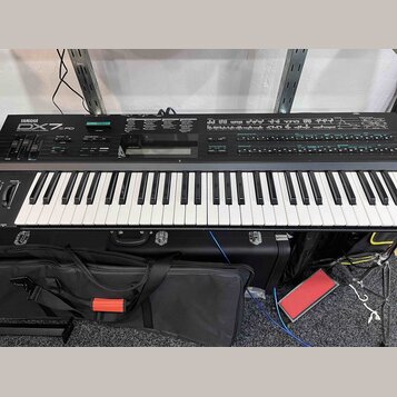 Černý digitální klavír s bílými a černými klávesami. Na levé straně je nápis YAMAHA DX7FD. Nástroj je umístěn v černém transportním kufru s kovovými…