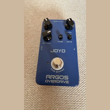 Modrý kytarový efekt s nápisy JOYO a ARGOS OVERDRIVE. Na horní straně jsou tři velké černé otočné knoflíky s označením ATTACK a LEVEL, dvě menší čern…