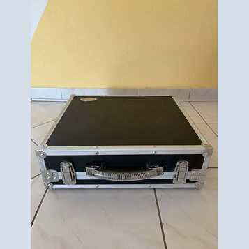 Černý transportní kufr s hliníkovými hranami a rohy. Na horní straně je nalepený oválný štítek s nápisem "DRUMCASE". Kufr má kovovou rukojeť a dva ko…