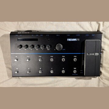 Černý kytarový procesor s nápisem FIREHAWK FX a logem LINE 6. Zařízení obsahuje displej, šest otočných ovladačů s popisky DRIVE, BASS, MID, TREBLE, F…