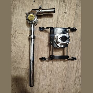 Na fotografii jsou dvě části chromovaného a černého kovového držáku na buben. Na horní části je žlutý oválný štítek s nápisem Pearl UNI-LOCK SYSTEM.…