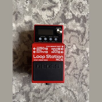 Červený kytarový pedál značky BOSS s nápisem Loop Station RC-5. Na horní části je displej, tři ovládací tlačítka s popisky RHYTHM, SETUP a MEMORY, ot…