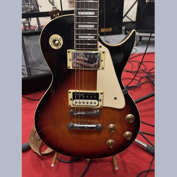 Elektrická kytara s tmelem v barvě sunburst (černá přecházející do hnědé). Má dva humbuckery s kovovými kryty, bílý pickguard a čtyři zlaté ovládací…