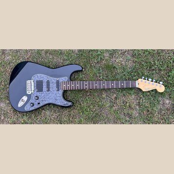 Černá elektrická kytara s šedým mramorovaným pickguardem a hnědým hmatníkem. Na hlavě je viditelné logo Fender a nápis STRATOCASTER, níže je drobný n…