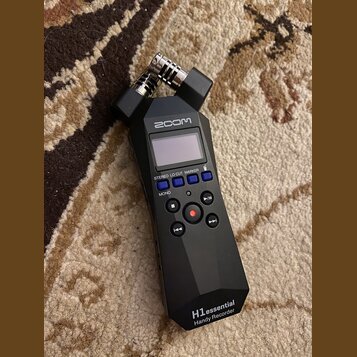 Černý digitální záznamový přístroj značky zoom. Na přední straně je displej, ovládací tlačítka a v dolní části nápis H1essential Handy Recorder. V ho…