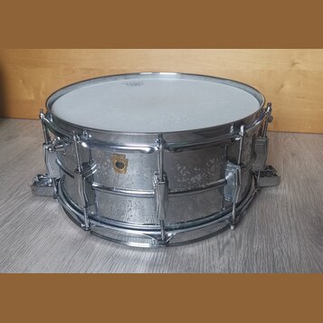 Kovový snare drum s chromovaným povrchem a bílým bubenovou skins. Na korpusu je viditelné zlaté logo ve tvaru štítu, ale text uvnitř něj není čitelný…