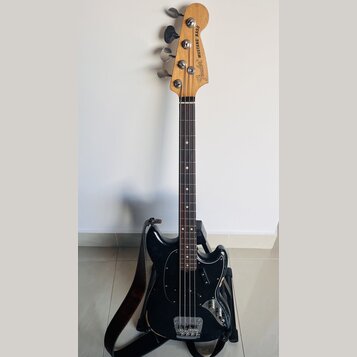 Černá elektrická basová kytara s přírodním dřevěným hmatníkem a hlavou. Na hlavě je nápis Fender MUSTANG BASS. Nástroj má černý plech ochranný kryt (…