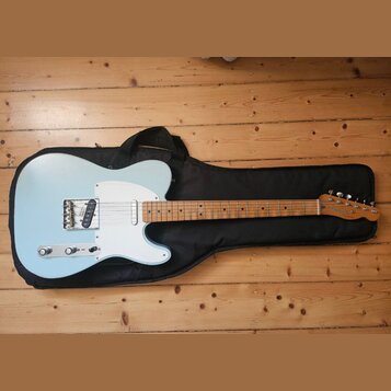 Světle modrá elektrická kytara typu Telecaster s bílým pickguardem, položená na černém softcasu. Na hlavě hředu je viditelné logo Fender a nápis MADE…