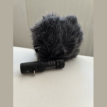 Černý mikrofon s nasazenou šedou chlupatou ochranou proti větru. Na ochraně je viditelné logo RØDE a na těle mikrofonu nápisy Videomic Me MADE IN AUS…