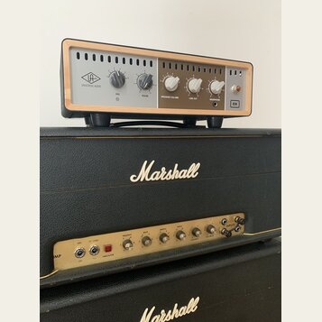 Na fotografii jsou dva kytarové zesilovače. Horní zařízení má stříbrný přední panel s dřevěným rámem, logo UNIVERSAL AUDIO a nápisy RIG, ROOM, SPEAKE…