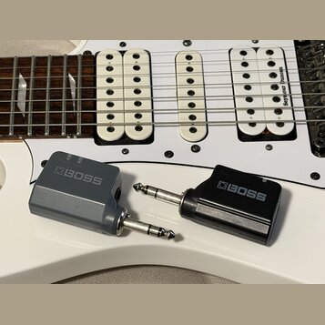 Na bílé elektrické kytáře leží dva šedé a černé adaptéry značky BOSS. Na bílém snímači kytary je viditelný nápis Seymour Duncan. Adaptéry mají kovové…