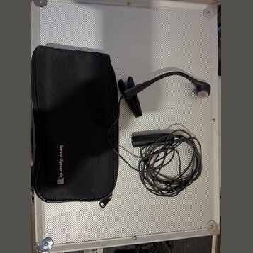 Černý mikrofon s ohebným hrdlem a připojeným kabelem. Vedle něj je černý přepravní pouzdro s bílým nápisem beyerdynamic))). Vše leží na stříbrném pov…