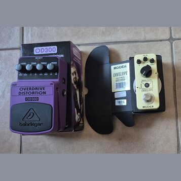 Na fotografii jsou dva kytarové efekty s krabicemi. Fialový pedál má nápisy OVERDRIVE DISTORTION, OD300 a logo behringer. Vedle něj je krémový pedál…