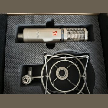 Stříbrný kondenzátorový mikrofon s mřížkou v horní části, umístěný v černé pěnové výplni. Na těle mikrofonu je červené logo s nápisem se a černý text…