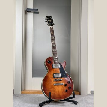 Elektrická kytara s tvarem singlecut a povrchovou úpravou v hnědém sunburstu. Na hlavě je viditelné logo Gibson. Kytara má dva humbuckery, čtyři otoč…