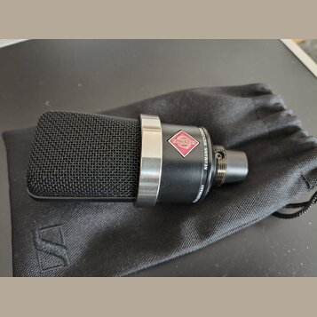 Černý kondenzátorový mikrofon s kovovou síťovkou a stříbrným prstencem, položený na černém látkovém vaku. Na těle mikrofonu je červenobílý kosočtverc…