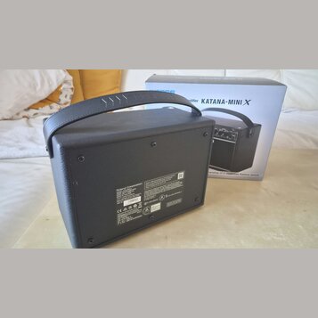 Černý kytarový kombo zesilovač s rukojetí, umístěný vedle původního kartónového obalu. Na krabici je nápis „amplifier KATANA-MINI X“. Na zadní straně…