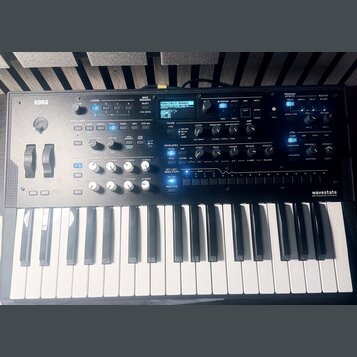 Šedý syntetizátor s klaviatura značky KORG, na jehož pravé straně je nápis wavestate. Přístroj disponuje displejem, několika posuvníky, otočnými knof…
