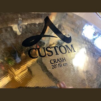 Detailní záběr na povrch kovového činely v zlatavé barvě. Na povrchu je černý nápis CUSTOM a CRASH 20"/51 cm spolu s grafickým logem ve tvaru stylizo…