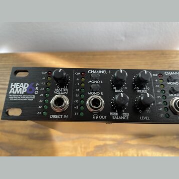 Černý rackový zesilovač sluchátek s nápisem HEAD AMP 6 PRO a popiskem PROFESSIONAL SIX CHANNEL HEADPHONE AMPLIFIER WITH EQ AND AUXILIARY MIXER. Na př…
