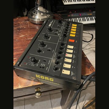 Černý kytarový syntetizér s nápisem KORG GUITAR SYNTHESIZER na spodní hraně. Na horním panelu jsou umístěny různé otočné potenciometry, přepínače a ž…
