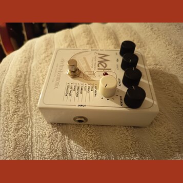 Bílý kytarový efekt s nápisem Electro-Harmonix NEW YORK CITY USA. Na horní straně je název modelu MeldaTone Case melody machine a seznam zvuků od 1 O…