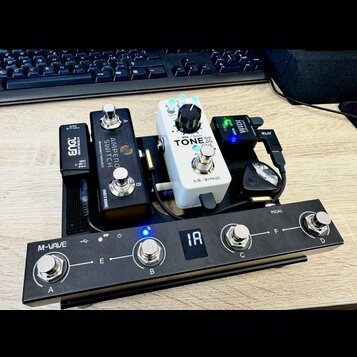 Sada kytarových efektů a ovládacích pedálů na černém podložkovém boardu. V popředí je černý pedalboard M-VAVE s čtyřmi spínači označenými A, B, C a D…