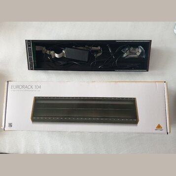 Fotografie zobrazuje černý Eurorack case s vloženým napájecím zdrojem a kabely, vedle jeho původního kartónového obalu. Na krabici je nápis EURORACK…