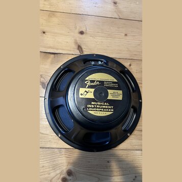Černý reproduktor s kovovým rámem, položený na dřevěném povrchu. Na zadní straně je žlutá nálepka s nápisy „SPECIALLY DESIGNED FOR Fender MUSICAL INS…