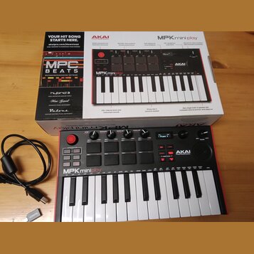 Na fotografii je kompaktní MIDI klaviatura AKAI PROFESSIONAL MPKminiplay v černé barvě s červeným okrajem, umístěná před svým původním kartonovým oba…