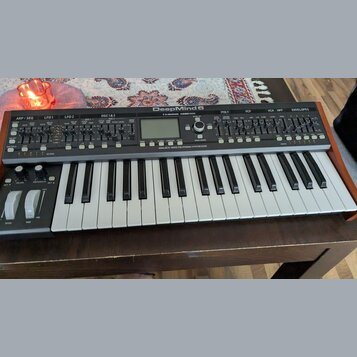 Šedý analogový syntetizátor s klaviaturaí a dřevěnými bočními panely. Na horním panelu je nápis DeepMind 6 a logo tc electronic. Pod ovládacími prvky…