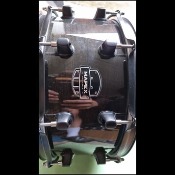 Černý buben s lesklým povrchem a kovovými obručmi. Uprostřed je umístěno logo s nápisem MAPEX a textem MPX MAPLE. Na povrchu jsou viditelné odrazy ok…