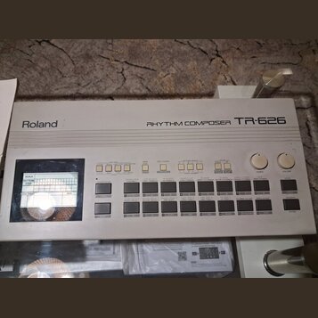 Bílý rytmický kompozér Roland RHYTHM COMPOSER TR-626. Zařízení obsahuje displej s mřížkou, několik řad ovládacích tlačítek a dva otočné ovladače ozna…