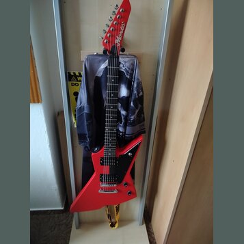 Červená elektrická kytara s ostrovním tvarem těla a hmatníkem z tmavého dřeva. Na hlavě je bílý nápis Aria. Kytara má dva humbuckery, ovládací knoflí…