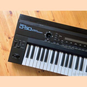 Černý syntetizátor s bílými a černými klávesami. Na horním panelu je nápis Roland D-50 LINEAR SYNTHESIZER. Jsou zde vidět různé ovládací tlačítka, po…