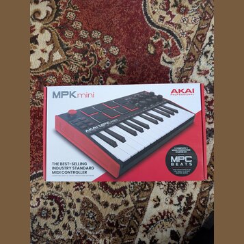 Fotografie zobrazuje krabici od MIDI kontroléru AKAI MPK mini. Na balení je zobrazen červený nástroj s klávesami a padem, doprovázený nápisy AKAI PRO…