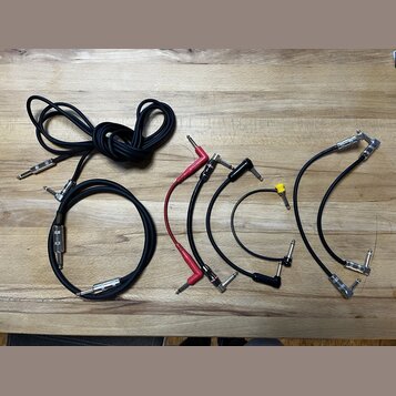 Sada různých audio kabelů s konektory typu jack položená na dřevěném stole. Je zde jeden dlouhý černý kabel, jeden kratší černý kabel s nápisem "KORD…