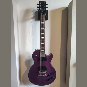 Fialová elektrická kytara s tvarem Les Paul, umístěná v nástěnním držáku. Na hlavě hředu je zlatý nápis Gibson. Kytara má dva humbuckery, čtyři ovlád…
