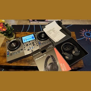 Na fotografii je černý DJ 컨트롤er značky Numark s modrým displejem a dvěma jogovými koly. Vedle něj leží krabice a černé sluchátka s nápisem Pioneer DJ…