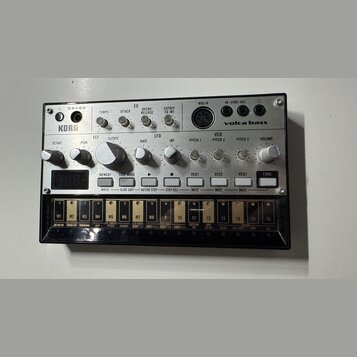 Stříbrný analogový syntetizátor s nápisy KORG a volca bass. Zařízení obsahuje řadu otočných knoflíků, tlačítek a digitální displej. V dolní části je…