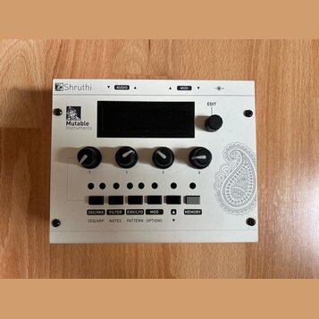 Bílý elektronický modul s nápisem Shruthi a logem Mutable Instruments. Zařízení má čtyři černé otočné ovladače označené čísly 1 až 4, pět malých zele…