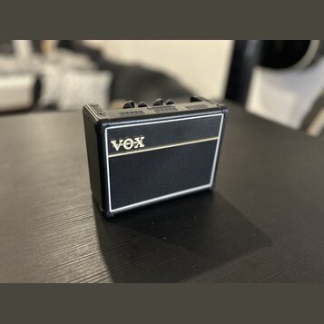 Malý kytarový zesilovač s černým povrchem z texturovaného materiálu a bílými lemovými pruhy. Na přední straně je umístěno zlaté logo VOX. Horní panel…