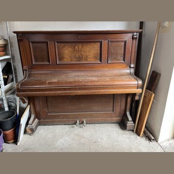 Hnědé dřevěné piano s výraznou masivní konstrukcí a dekorativními panely. Povrch je pokryt prachem a vykazuje známky opotřebení v podobě škrábanců. N…