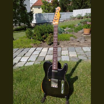 Elektrická kytara s černým bodies, hnědým hnedem na okrajích a tmavým hmatníkem. Na hlavě je viditelné logo Fender Telecaster. Kytara má černý pickgu…